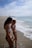Two women in swimsuits stand at the edge of a sandy beach, with gentle waves approaching their feet. The sky is partly cloudy, and a distant structure is visible along the shoreline.