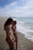 Two women in swimsuits stand at the edge of a sandy beach, with gentle waves approaching their feet. The sky is partly cloudy, and a distant structure is visible along the shoreline.
