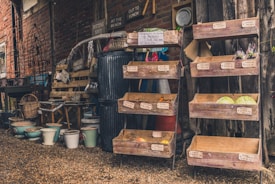 A rustic outdoor market stall displays wooden crates filled with various fruits and vegetables. Handwritten signs with prices are attached to each crate. There are several stacked empty pots of various sizes on the ground. A large trash bin and an assortment of gardening tools are visible. The background is a brick wall with wooden elements, and a small clock is mounted on the wall.