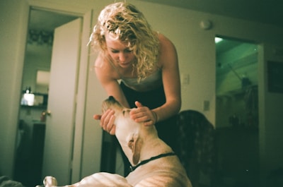 A person with curly blonde hair is leaning over and gently holding the face of a light-colored dog. The setting appears to be indoors, possibly a living room, with an open door leading to an adjacent room. Soft lighting creates a warm and intimate atmosphere.