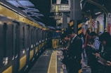A group of people stand on a train platform, many looking at their phones. A yellow train is stopped at the platform, ready to depart. The scene appears busy and urban, with overhead signs in Japanese indicating the location. The platform has yellow safety lines and barriers alongside the tracks.