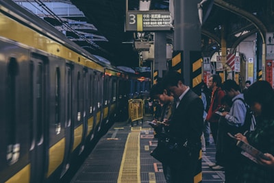 A group of people stand on a train platform, many looking at their phones. A yellow train is stopped at the platform, ready to depart. The scene appears busy and urban, with overhead signs in Japanese indicating the location. The platform has yellow safety lines and barriers alongside the tracks.