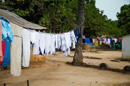 A rural outdoor setting with clothes hanging on multiple clotheslines stretching across the scene. The clothes are mostly white, though there are some colorful garments further along the line. The lines are strung between a tree and what appears to be a small building. Surrounding the area are trees with dense green foliage. The ground is unpaved, and there are modest structures in the background.