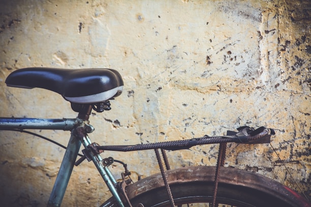An old bicycle seat attached to a weathered bike frame is positioned against a textured, worn-out yellow wall. The bike's metal appears rusty and paint-chipped, with a black rack situated over the rear wheel.