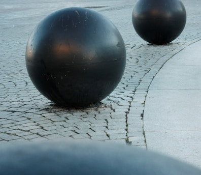 Large black spherical objects placed on a cobblestone surface, with a curved path of smooth stone tiles visible nearby. The setting appears to be outdoors.