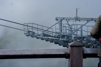 A metal structure with a complex assembly of cables and pulleys is set against a foggy backdrop. Partial view of a person wearing a jacket is visible in the foreground.