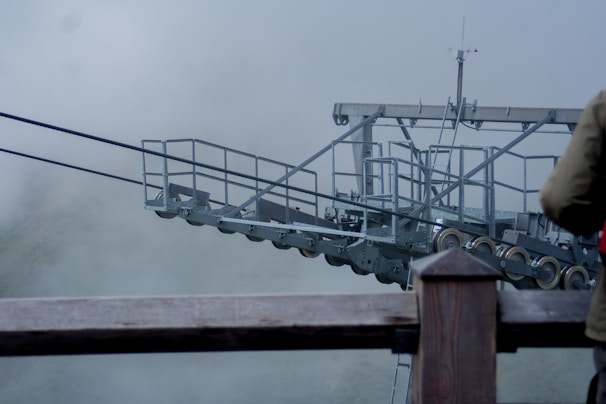 A metal structure with a complex assembly of cables and pulleys is set against a foggy backdrop. Partial view of a person wearing a jacket is visible in the foreground.