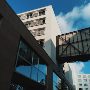 A modern, multi-story building showcasing large glass windows and a connecting walkway between structures. The architecture features clean geometric lines, and shadows are cast on the facade, creating a dynamic visual effect against a bright blue sky.