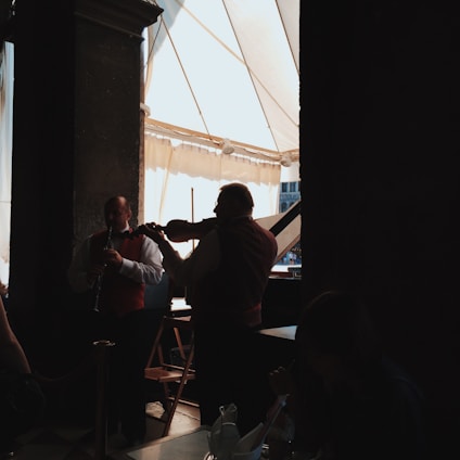 Musicians are playing instruments in a dimly lit indoor setting with large windows allowing natural light to filter in. The focus is on a violinist and another musician, both wearing dark vests over white shirts, performing together.