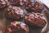 Chocolate cupcakes topped with colorful sprinkles arranged on a tray. The icing is glossy and creamy, adding a rich texture to the overall appearance.