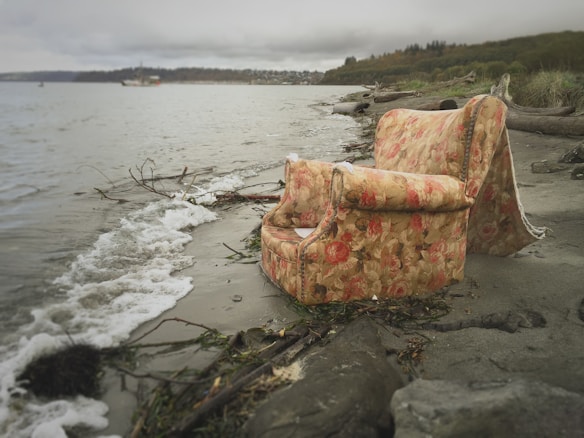 An abandoned floral-patterned armchair and sofa sit partially submerged on a gloomy, rocky shoreline. The ocean waves gently lap at the furniture, surrounded by wet sand, patches of seaweed, and driftwood. The sky is overcast, casting a muted light over the scene, while distant trees and a faintly visible boat are seen on the horizon.