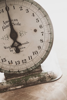An old mechanical weighing scale with a rustic appearance. The dial has numbers from 0 to 23 and the text 'American Family Scale' printed on it. The base shows signs of wear with chipped paint and a greenish hue.