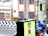 Two pigeons are perched on a rectangular box with a radiation warning sign. The background includes a cityscape with buildings and a road featuring a black and white chevron pattern on the barriers. Blurred figures suggest motion, likely pedestrians or vehicles.