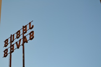 A metal sign with the letters oriented vertically against a clear blue sky. The letters appear to be arranged in an industrial style, attached to tall poles, suggesting a retro or vintage aesthetic.