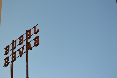 A metal sign with the letters oriented vertically against a clear blue sky. The letters appear to be arranged in an industrial style, attached to tall poles, suggesting a retro or vintage aesthetic.