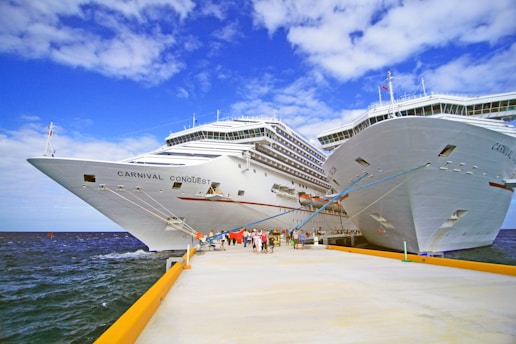 Two large cruise ships named Carnival Conquest docked side by side at a pier. The scene is bustling with people walking along the pier under a sky filled with scattered clouds.