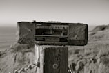 A vintage Sony radio cassette recorder is mounted on a wooden post outdoors. The background includes a blurred view of the ocean and natural landscape, with grass and hills. The photo has a sepia tone, giving it an aged appearance.