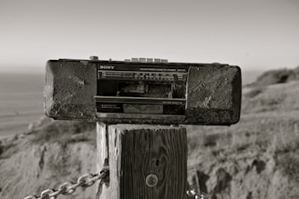 A vintage Sony radio cassette recorder is mounted on a wooden post outdoors. The background includes a blurred view of the ocean and natural landscape, with grass and hills. The photo has a sepia tone, giving it an aged appearance.