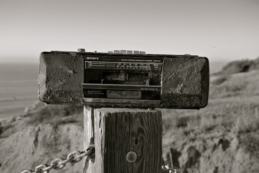 A vintage Sony radio cassette recorder is mounted on a wooden post outdoors. The background includes a blurred view of the ocean and natural landscape, with grass and hills. The photo has a sepia tone, giving it an aged appearance.