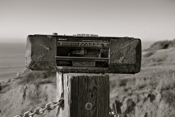 A vintage Sony radio cassette recorder is mounted on a wooden post outdoors. The background includes a blurred view of the ocean and natural landscape, with grass and hills. The photo has a sepia tone, giving it an aged appearance.