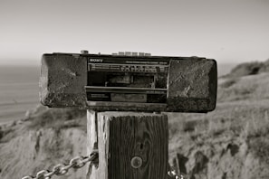 A vintage Sony radio cassette recorder is mounted on a wooden post outdoors. The background includes a blurred view of the ocean and natural landscape, with grass and hills. The photo has a sepia tone, giving it an aged appearance.