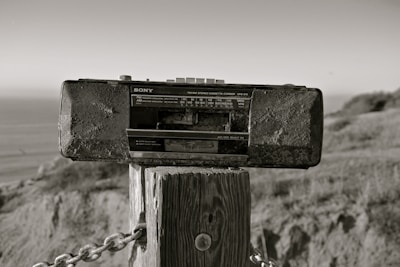 A vintage Sony radio cassette recorder is mounted on a wooden post outdoors. The background includes a blurred view of the ocean and natural landscape, with grass and hills. The photo has a sepia tone, giving it an aged appearance.