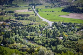 A lush, green landscape with a winding road cutting through a small town featuring dense tree coverage and scattered houses. The surrounding fields and hills create a serene, expansive view with abundant greenery.