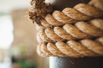 A close-up view of a thick, coiled rope with a definitive twisted pattern. The texture of the fibers is prominent, giving it a rugged and natural appearance. The background is blurred, drawing attention primarily to the rope.
