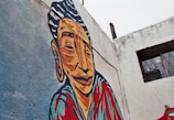 A vibrant mural painted on a weathered concrete wall features a stylized portrait of a person with bold lines and exaggerated features. The figure is depicted with a serene expression and adorned in colorful clothing. The background consists of a light blue texture, while part of the wall displays a window with metal bars.