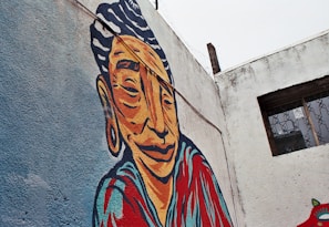 A vibrant mural painted on a weathered concrete wall features a stylized portrait of a person with bold lines and exaggerated features. The figure is depicted with a serene expression and adorned in colorful clothing. The background consists of a light blue texture, while part of the wall displays a window with metal bars.