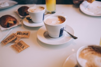 Several cups of coffee with frothy tops are placed on a white table. There are packets of sugar labeled 'BREK' and 'VELOCE' scattered on the table along with some pastries, possibly croissants. In the background, there is a glass of orange juice and another plate with a croissant being held by a person.