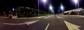 A wide road at night with bright streetlights illuminating the area. There are arrows painted on the road, indicating traffic directions. To the left, a person in an orange top is sitting on the curb. On either side of the road, covered walkways and greenery are visible.