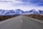 An open road stretches towards a range of snow-capped mountains under a partly cloudy sky. The road is flanked by dry bushes and shrubs, indicating a semi-arid environment, and the perspective is centered, inviting a sense of journey and adventure.