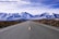 An open road stretches towards a range of snow-capped mountains under a partly cloudy sky. The road is flanked by dry bushes and shrubs, indicating a semi-arid environment, and the perspective is centered, inviting a sense of journey and adventure.