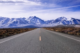 An open road stretches towards a range of snow-capped mountains under a partly cloudy sky. The road is flanked by dry bushes and shrubs, indicating a semi-arid environment, and the perspective is centered, inviting a sense of journey and adventure.