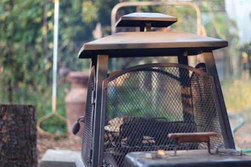 A rustic metal outdoor fireplace with a grid pattern on its door is situated in a garden setting. The background is blurred, featuring greenery and garden implements. The light creates a soft, natural ambiance, highlighting the texture of the metal and surrounding vegetation.