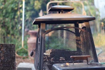 A rustic metal outdoor fireplace with a grid pattern on its door is situated in a garden setting. The background is blurred, featuring greenery and garden implements. The light creates a soft, natural ambiance, highlighting the texture of the metal and surrounding vegetation.