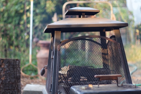 A rustic metal outdoor fireplace with a grid pattern on its door is situated in a garden setting. The background is blurred, featuring greenery and garden implements. The light creates a soft, natural ambiance, highlighting the texture of the metal and surrounding vegetation.