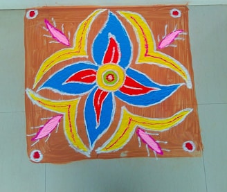 A colorful rangoli design featuring a central floral pattern with spiraling petals. The design incorporates vibrant shades of blue, red, yellow, and pink, accented with white outlines. The background is a rustic brown color, and the pattern is neatly framed on a tiled floor.