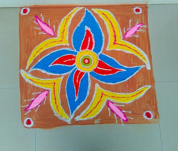 A colorful rangoli design featuring a central floral pattern with spiraling petals. The design incorporates vibrant shades of blue, red, yellow, and pink, accented with white outlines. The background is a rustic brown color, and the pattern is neatly framed on a tiled floor.