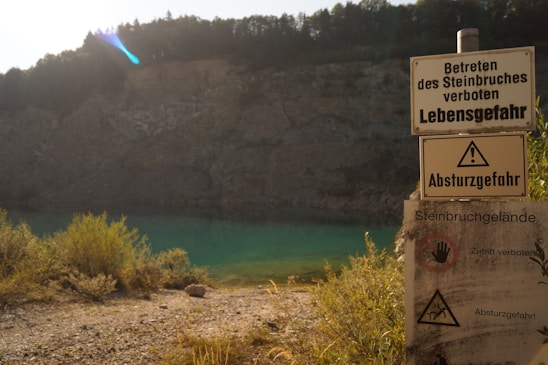 A rocky landscape with a quarry in the background, featuring a turquoise water body. Various warning signs with messages in German indicating danger and prohibition of entry are prominently displayed.