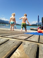 Two children wearing swimsuits are walking on a wooden dock towards a scenic lake under a clear blue sky. In the background, there are mountains and a few clouds. A blue towel and an orange inflatable toy are visible on the dock.