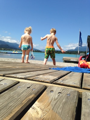Two children wearing swimsuits are walking on a wooden dock towards a scenic lake under a clear blue sky. In the background, there are mountains and a few clouds. A blue towel and an orange inflatable toy are visible on the dock.