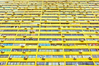 A large residential building featuring numerous identical windows arranged in a precise grid pattern. The exterior is predominantly yellow, and each window displays varied curtains and objects. Air conditioning units are mounted outside many of the windows.