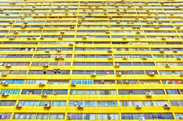 A large residential building featuring numerous identical windows arranged in a precise grid pattern. The exterior is predominantly yellow, and each window displays varied curtains and objects. Air conditioning units are mounted outside many of the windows.