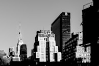 A monochrome view of a cityscape featuring several high-rise buildings. Among them, a prominent structure has the words 'NEW YORKER' prominently displayed on top. The skyline includes an iconic skyscraper with a spire, possibly the Empire State Building. Shadows and light create contrast on the surfaces of the structures.