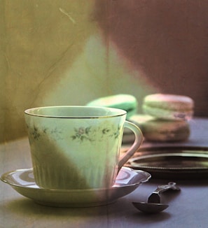 A delicate porcelain teacup with a floral pattern sits on a matching saucer. In the background, there are soft-focus macarons stacked near a mirror. A small antique spoon lies nearby, adding to the vintage feel of the setting.
