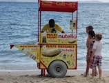 A beachside snack cart with vibrant advertisements for Nutella beignets and cold drinks. A vendor, wearing a yellow shirt, is serving three children who are waiting eagerly. The cart is positioned on the sandy beach near the water's edge, with the ocean visible in the background.
