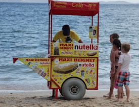 A beachside snack cart with vibrant advertisements for Nutella beignets and cold drinks. A vendor, wearing a yellow shirt, is serving three children who are waiting eagerly. The cart is positioned on the sandy beach near the water's edge, with the ocean visible in the background.