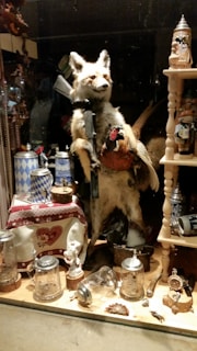A taxidermy fox is posed upright with a rifle and a pheasant in its hands. Surrounding the fox are various decorative items, including beer steins with Bavarian designs, glass mugs, and ornate metal objects. The scene resembles an eclectic display of rustic and hunting-themed memorabilia with a playful twist.